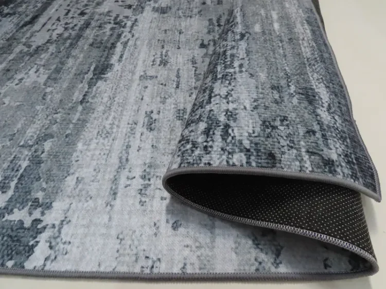 A rug with grey tones and a textured pattern. The edge of the rug is folded over, revealing a black non-slip backing