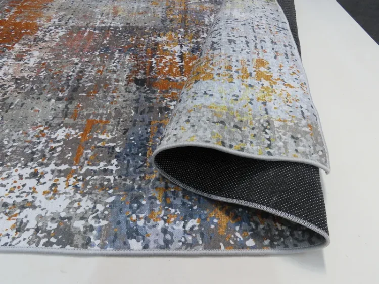 A rug with a modern, abstract design featuring splashes of grey, orange, yellow, and white colors. The rug is partially rolled up, revealing its black, non-slip backing