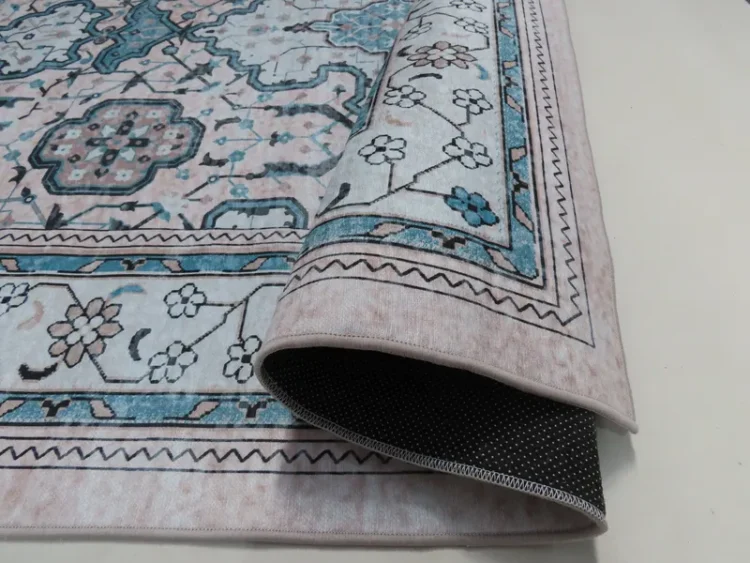 A rug with a detailed design with intricate floral and geometric patterns in blue and beige. The edge of the rug is rolled to show the backside, revealing a dotted non-slip surface