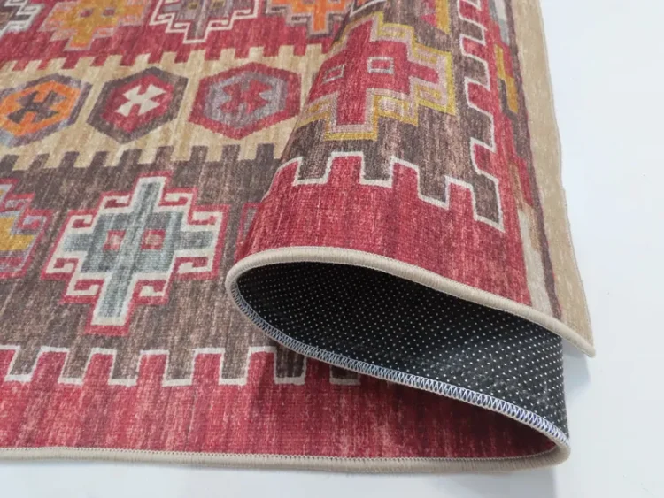 A rug with a vibrant pattern with geometric shapes in red, orange, and yellow hues. The close-up image shows the rug partially rolled, revealing a black dotted underside