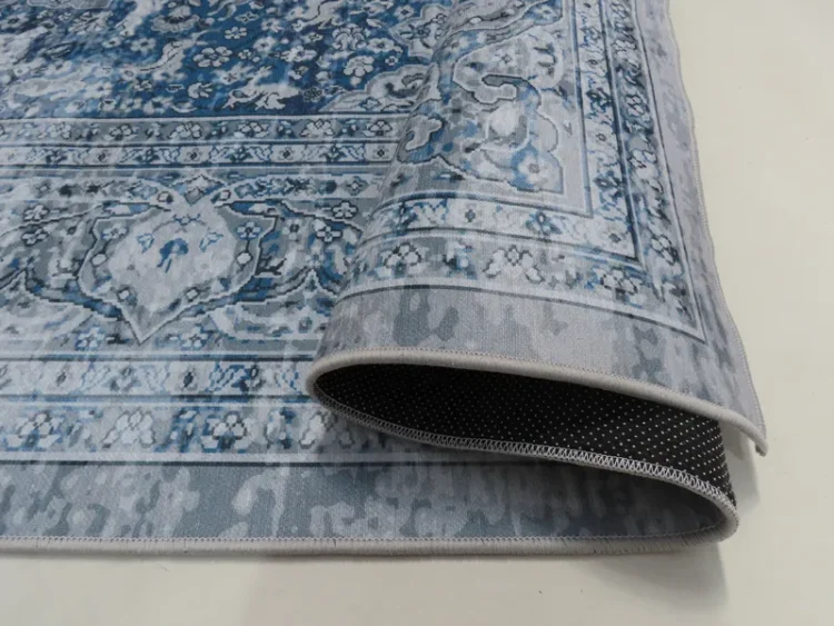 A rolled edge of a rug with an intricate floral pattern in grey and blue tones. The back of the rug has a non-slip black backing with small white dots