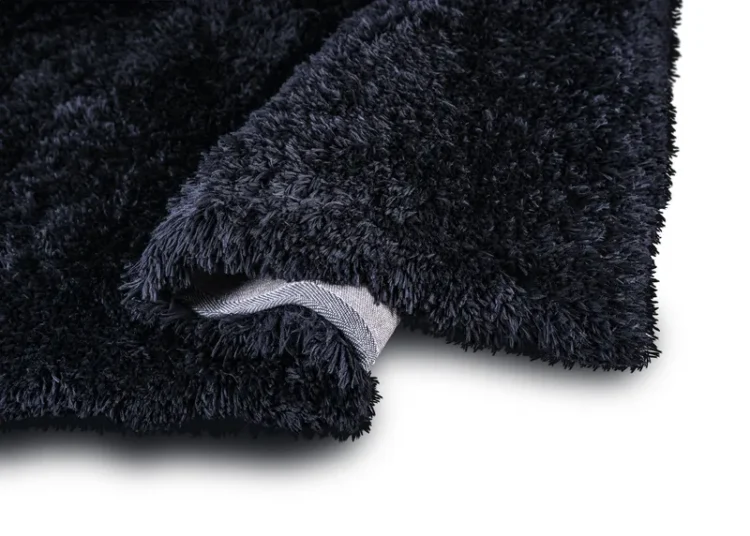 A plush, shaggy black rug, revealing its thick, soft texture. The rug is slightly folded, exposing a bit of its underside, which is a light grey woven material