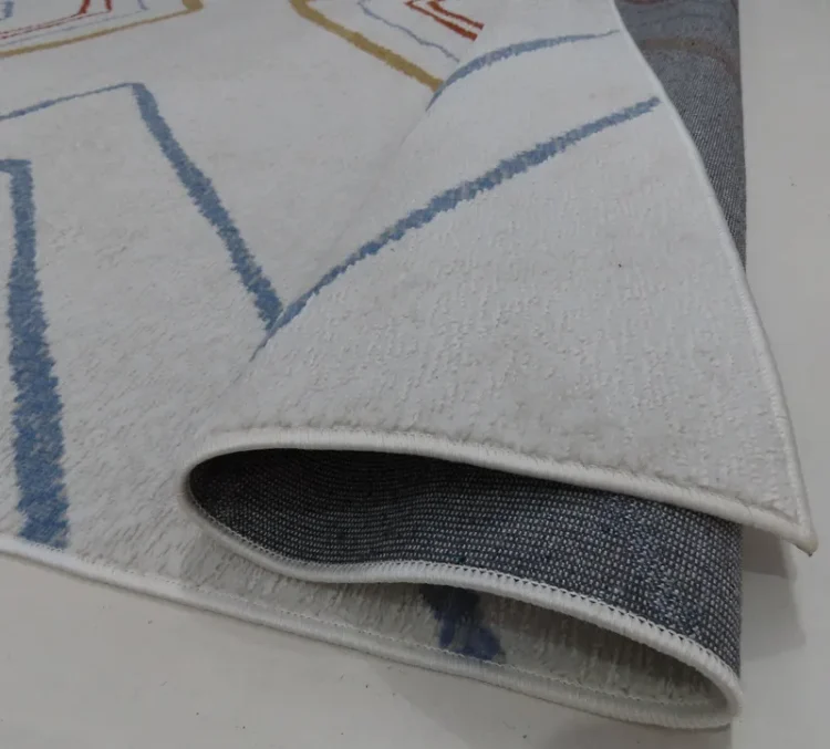 A cream-colored rug with blue and gold lines forming a diamond pattern. The rug is partially folded to reveal a textured, dark backing