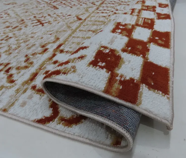 A beige and light blue geometric-patterned rug with one edge lifted to reveal the underside. The rug has a grey backing with a small dotted pattern