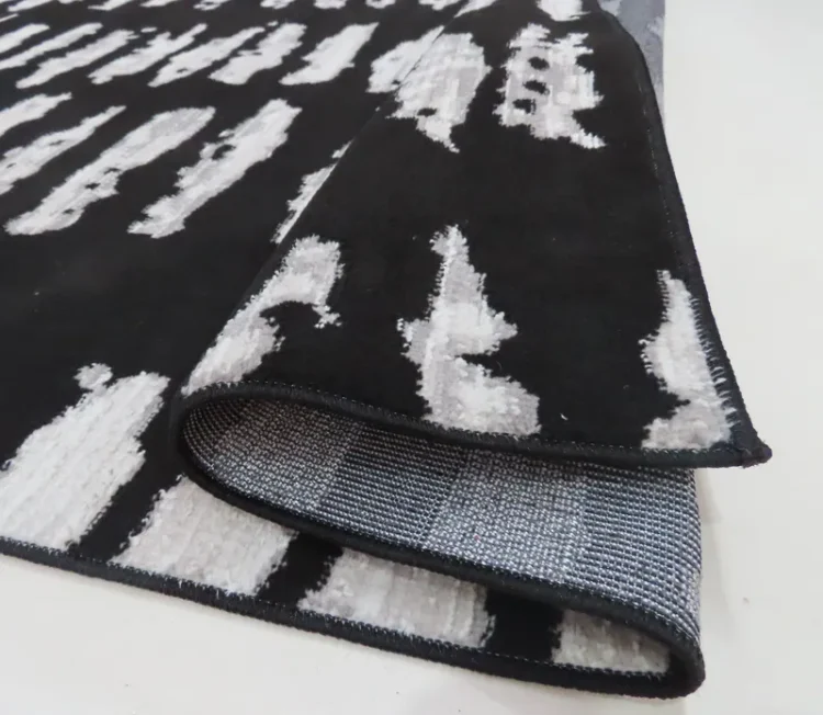 A black rug with irregular white horizontal stripes arranged in vertical columns. The edge of the rug is lifted to reveal a grey backing with a grid pattern, and the rug has black stitching along the edges