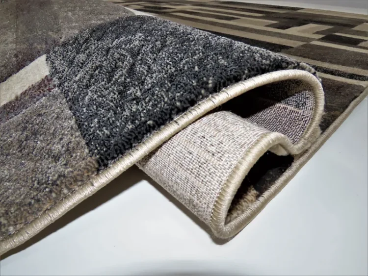 A rug with a geometric pattern in shades of brown, black, and cream. The edge of the rug is folded to show the backside, which has a simple woven texture