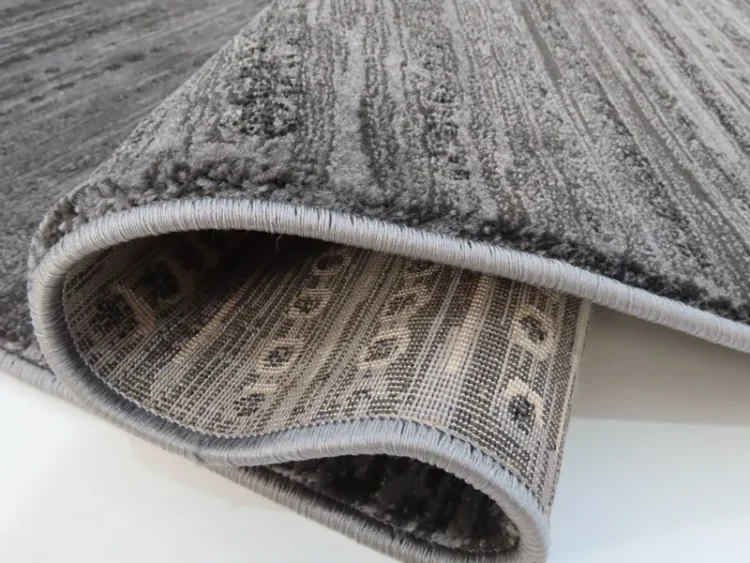 A rug with a dark grey colour with a subtle striped pattern and a soft, plush texture. The edge of the rug is rolled to show the backside, which has a woven, patterned fabric