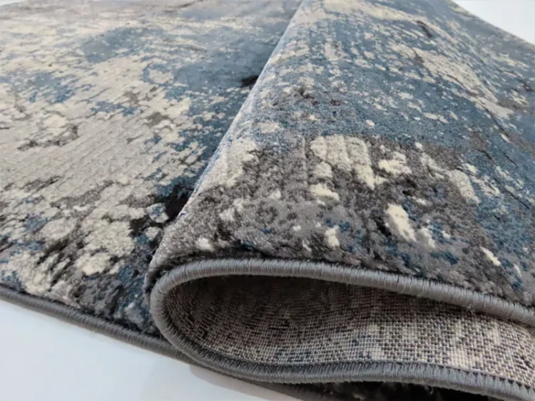 A folded rug, showing its edge and part of the underside. It has a dark blue and grey abstract pattern with a textured, splattered design