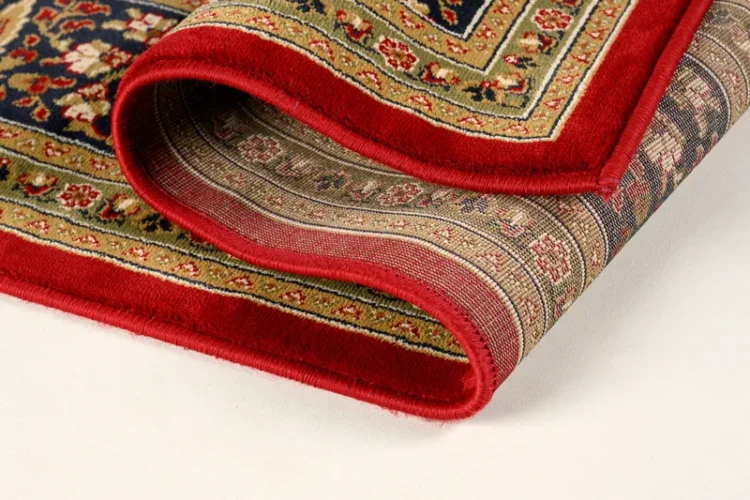 A red rug with folded edges. The rug has a floral border with intricate designs in green, beige, and gold