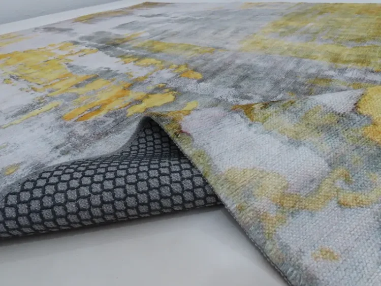 A rug with an abstract design in grey and yellow tones. The edge of the rug is lifted, revealing a grey backing with a hexagonal pattern.