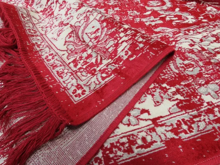 A traditional red and cream floral-patterned rug with the edge lifted to reveal the underside. The rug features red fringes along the edges