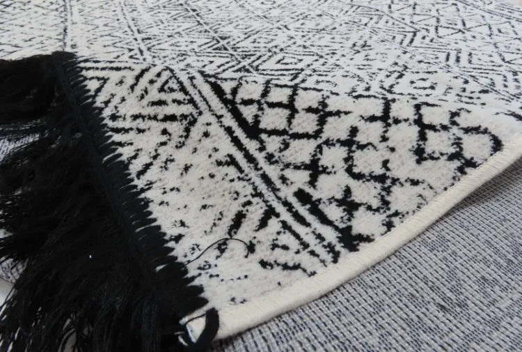 A black and white geometric-patterned rug with intricate designs, including diamonds and triangles. One edge of the rug is lifted to reveal the underside, and the rug features black fringes along the edge